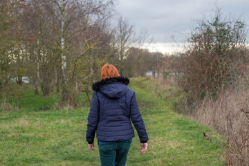 Naklejka na ściany i meble Middle aged woman with red hair seen walking away from the camera, along a nature trail in a popular nature reserve. She is seen wearing a winter jacket and attire.