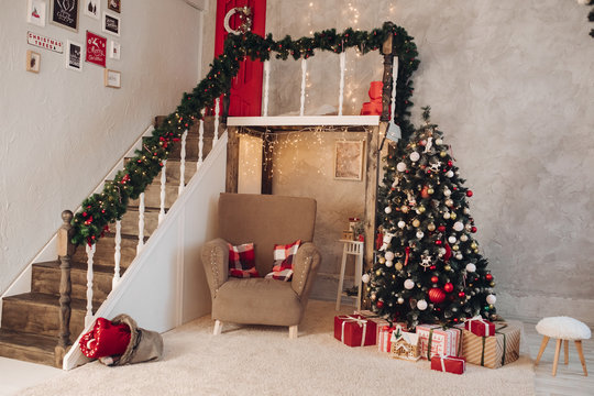 Stock photo of beautifully decorated living room with cozy armchair, Christmas tree and stairs with tinsel and garland. Cozy Christmas room with wrapped gifts under the tree.