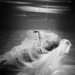 Black and white image. Underwater photo beautiful blonde wearing in white flying dress, swimming in pool underwater. Copy space.