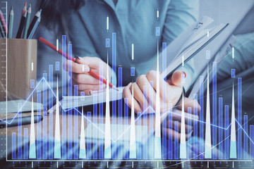 A woman hands writing information about stock market in notepad. Forex chart holograms in front....