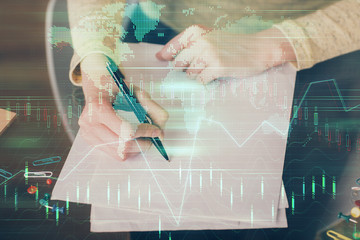 Hand taking notes in notepad. Forex chart holograms in front. Concept of research. Multi exposure