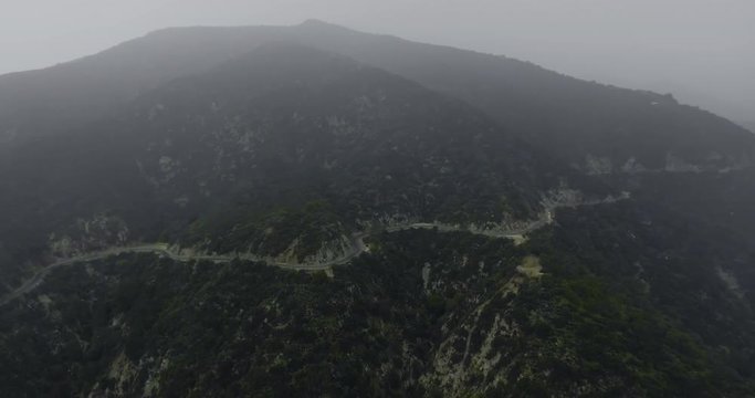 Drone shot dark mountains with a dense forest and a winding road under a foggy shroud (Santa Anita Canyon, California, USA)
