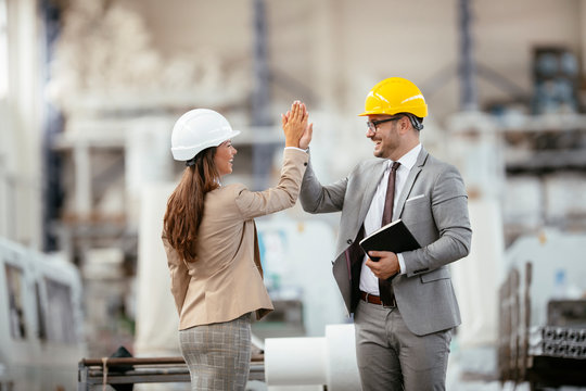 Businessman and businesswoman in factory. Man and woman in suits with helmets in factory giving each other high five.