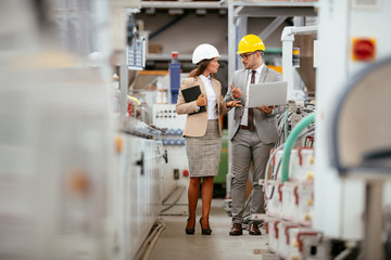 Fototapeta premium Businessman and businesswoman in factory. Man and woman in suits with helmets in factory discussing work.
