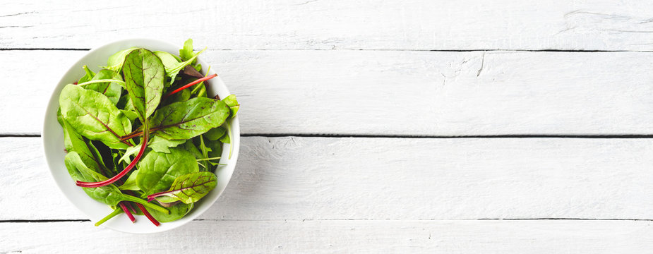 Overhead Shot Of Fresh Green Salad With Spinach, Arugula And Beetroot Leaves In Bowl On White Wooden Background With Copyspace. Banner