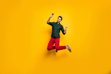 Full length body size photo of cheerful positive guy showing v-sign taking selfie wearing green t-shirt red pants trousers smiling toothily jumping up isolated vivid color background