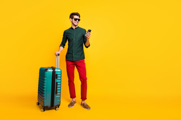 Full size profile side photo of cheerful traveler guy on summer vacation reach destination hold package use cellphone reserve taxi wear green shirt pants trousers isolated yellow color background