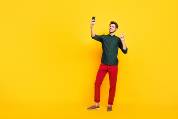 Full body photo of positive cheerful guy have fun spring holidays tale selfie on his smartphone make v-signs greet blog followers wear casual style outfit sneakers isolated yellow color background