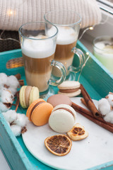 Two cups of hot coffee with milk on turquoise tray near the window. Macaroons, dried orange peels, cinnamon rolls and cotton flowers on marble plate. Knitted sweater and string lights on background