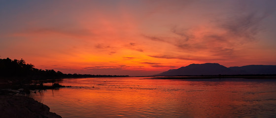 Panoramic view of sunset on the african Zambezi river. The dramatic, red sky reflects on the surface of the border river. View over the flood plain to the mountains on the Zambian side of the river.