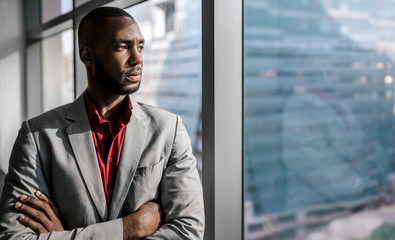 Confident black african businessman standing with his arms crossed looking out the office window in...