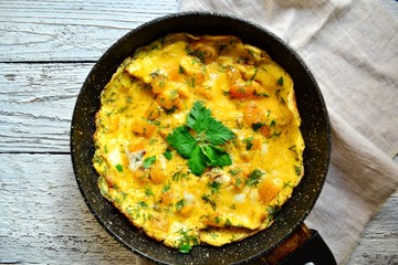 Appetizing omelet on a wooden light background. Omelet with yellow tomato, sweet yellow pepper and parsley leaves in a pan. Tasty breakfast.