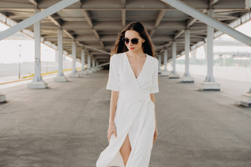 Obraz premium Portrait of a young woman in sunglasses. The girl in the white dress. Walk around the city. Long-haired brunette. Female portrait. Beauty and fashion. Gray background. Bridges and buildings. Photomode