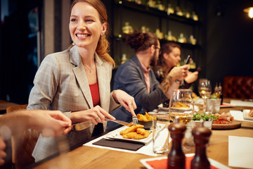Beautiful caucasian smiling woman cutting food, chatting with friends and smiling. Restaurant interior.