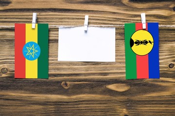 Hanging flags of Ethiopia and New Caledonia attached to rope with clothes pins with copy space on white note paper on wooden background.Diplomatic relations between countries.