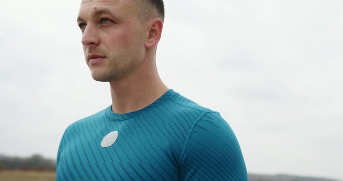 Revolving view of an attractive man in blue sportswear looking down then lifting his head as the camera pans around him in a close up portrait against cloudy sky