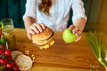 Beautiful young woman decides eating hamburger or apple in kitchen. Cheap junk food vs healthy diet