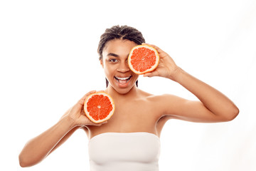 Beauty Concept. Young african woman wearing top isolated on white covering eye with grapefruit half mouth opened playful