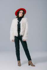 elegant trendy girl posing in white fur coat and red hat on grey