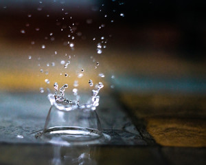Raindrop splashing into puddle on tile. Yellow and gray colors with reflections seen in background. 
