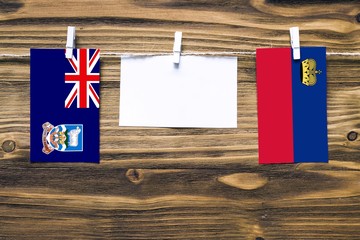 Hanging flags of Falkland Islands and Liechtenstein attached to rope with clothes pins with copy space on white note paper on wooden background.Diplomatic relations between countries.