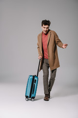 happy stylish man in beige coat dancing with travel bag on grey
