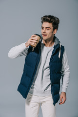 happy handsome man in warm waistcoat drinking coffee to go, isolated on grey