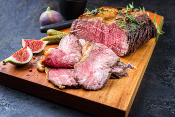 Traditional lunch meat with sliced cold cuts roast beef with gherkin and fig fruit as closeup on a modern design cutting board