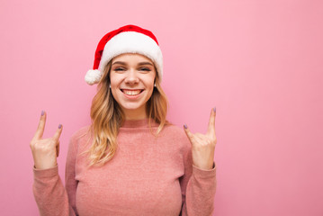 Portrait of happy lady in christmas hat listening to music in headphones and showing hands gesture Rock and pink background, looking at camera and smiling. Isolated.