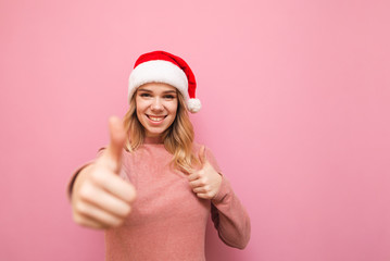 Happy blonde girl in santa hat and pink sweater stands on pink background and shows thumbs up, looks at camera and smiles. Smiling lady in santa hood shows gesture like. Isolated. X-mas