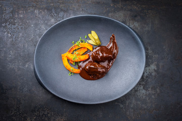 Traditional German braised beef cheeks in brown red wine sauce with pumpkin slices and herbs offered as closeup in a modern design plate with copy space