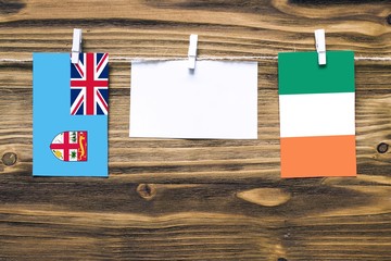 Hanging flags of Fiji and Ireland attached to rope with clothes pins with copy space on white note paper on wooden background.Diplomatic relations between countries.