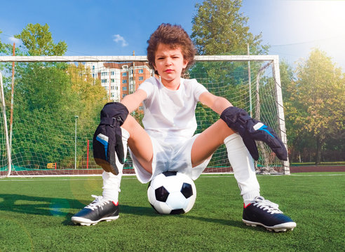 Thoughtful little boy sitting on the soccer ball against the goal on a sportsfield.Boy goalkeeper in football sportswear on stadium with ball.Pose of the winner.Confidence and the will to win concept.
