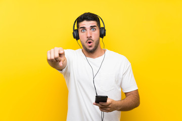 Young handsome man listening music with a mobile over isolated yellow wall surprised and pointing front