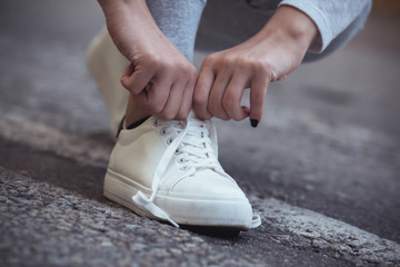 Naklejka premium girl squatted down to tie shoelaces on white sneakers on asphalt road, autumn sport concept outdoors