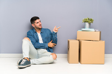 Handsome young man moving in new home among boxes pointing finger to the side