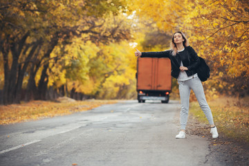 young woman stands and waits on an asphalt road in cold autumn weather, a girl tries to get from a suburban highway by a hitchhiker showing a hand gesture