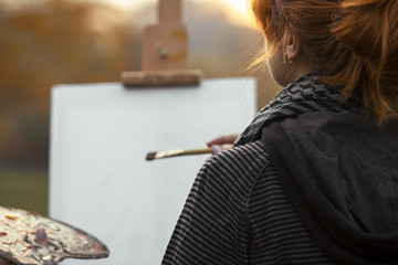 back head of red-haired girl with a bun painting a picture on an easel in nature, a young woman engaged in art and enjoying beautiful landscape at sunset ,concept hobby and lifestyle