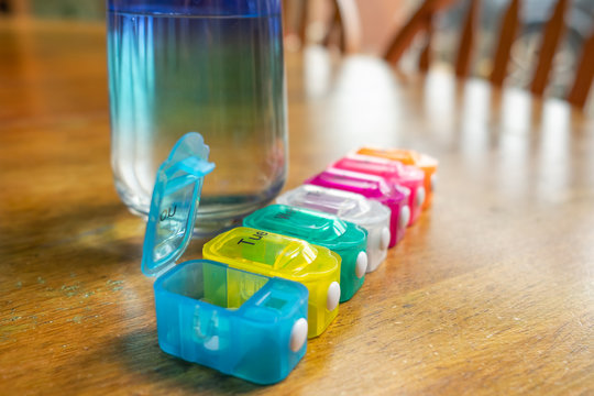 Shallow focus of a plastic pill dispenser seen on stable next to a glass of water. Each compartment holds the tablets and pills to be taken by the patient on that day.