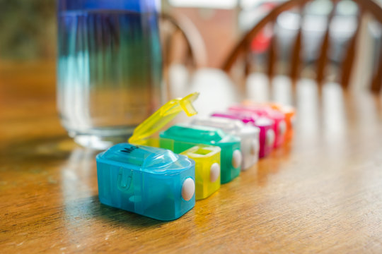 Shallow focus of a plastic pill dispenser seen on stable next to a glass of water. Each compartment holds the tablets and pills to be taken by the patient on that day.