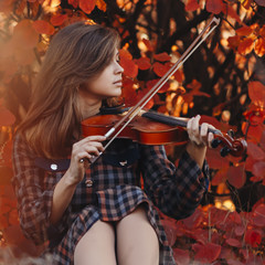 Fototapeta premium beautiful young woman sitting on the ground with a violin on a background of red bush, girl in dress leading the bow along the strings of musical instrument in nature, a concept of hobby and art