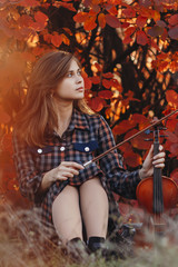 Obraz na płótnie Canvas young woman sitting on the ground with a violin on a background of red autumn foliage, romantic girl in dress with a musical instrument on nature, a concept of hobby and art