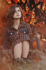 beautiful young woman sitting on the ground with a violin on a background of red foliage, romantic girl in dress inspired by nature for creative activity, concept of hobby and music