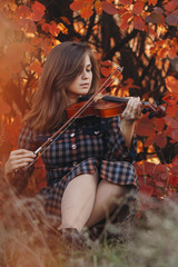 Obraz na płótnie Canvas autumn portrait of beautiful woman sitting on the ground with a violin under chin on a background of red foliage, girl enjoying sound of musical instrument, a concept of passion in art