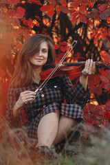 Obraz na płótnie Canvas autumn portrait of beautiful woman sitting on the ground with a violin under chin on a background of red foliage, girl engaged in playing a musical instrument in nature, a concept of passion in art
