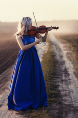 woman in long dress playing violin on background of field path with a club of dust, girl engaged in...