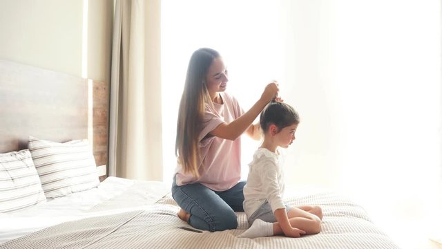 Beautiful charming woman dressed in casual clothes spending time with little daughter, holding long hair, putting scrunchy, morning time concept, family portrait