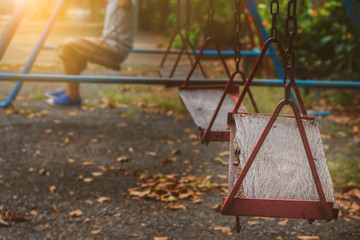 Blurred alone boy sitting on old wooden garden swings in abandoned playground with sunset lighting background, Selective focus at the front wood seat.