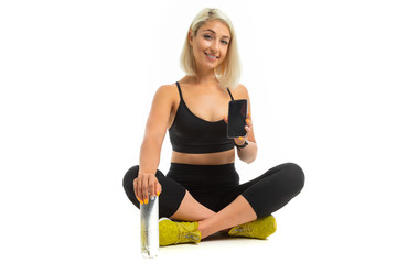 Beauiful sport caucasian girl sits on floor, cross legs, shows a mobile phone and keeps a bottle of water isolated on white background