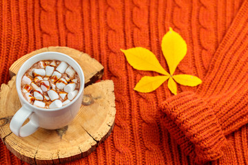 White ceramic cup with a hot cocoa drink decorated on top with chocolate topping stands on a round wooden stand. Yellow chestnut sheet. A red sweater of large knitting as a back background. Copyspace